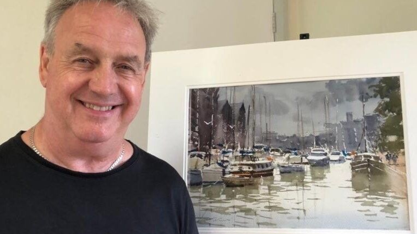 Smiling artist standing beside a framed painting of sailboats in a marina.