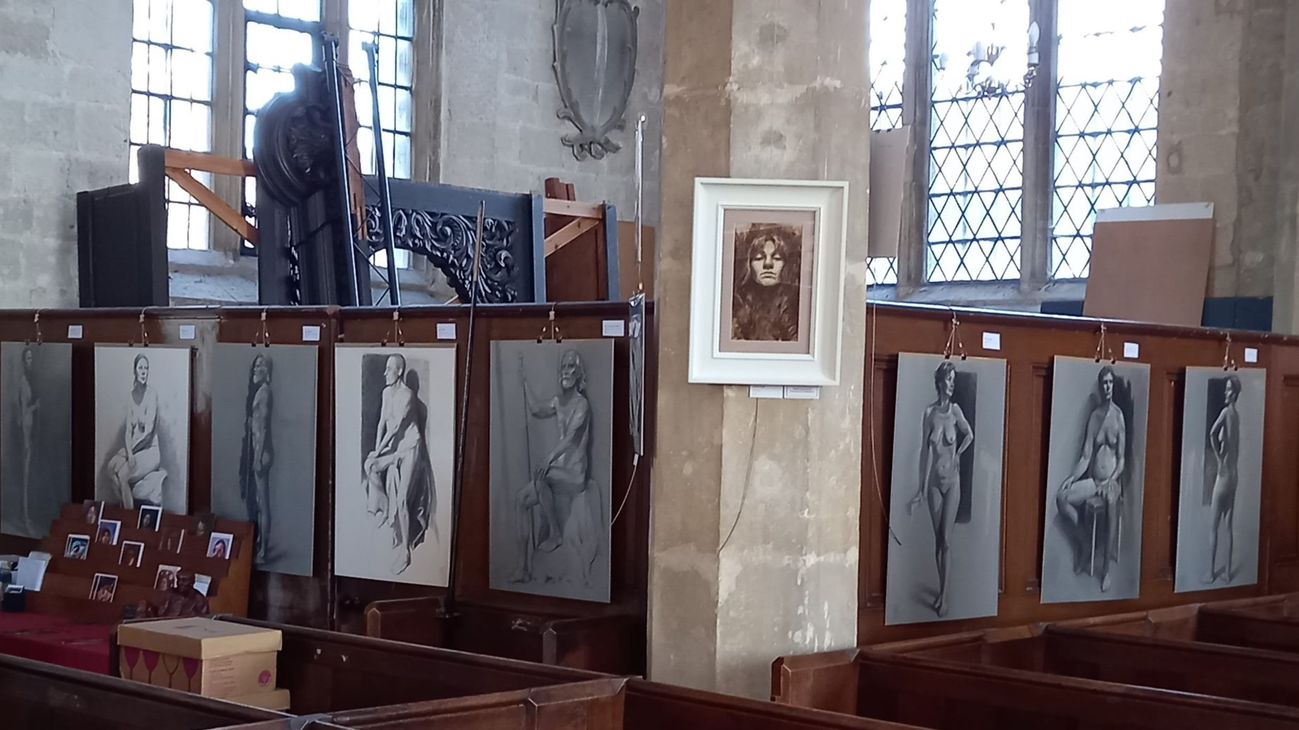 Art exhibition in a church with framed black-and-white drawings displayed on wooden pews and stone columns.