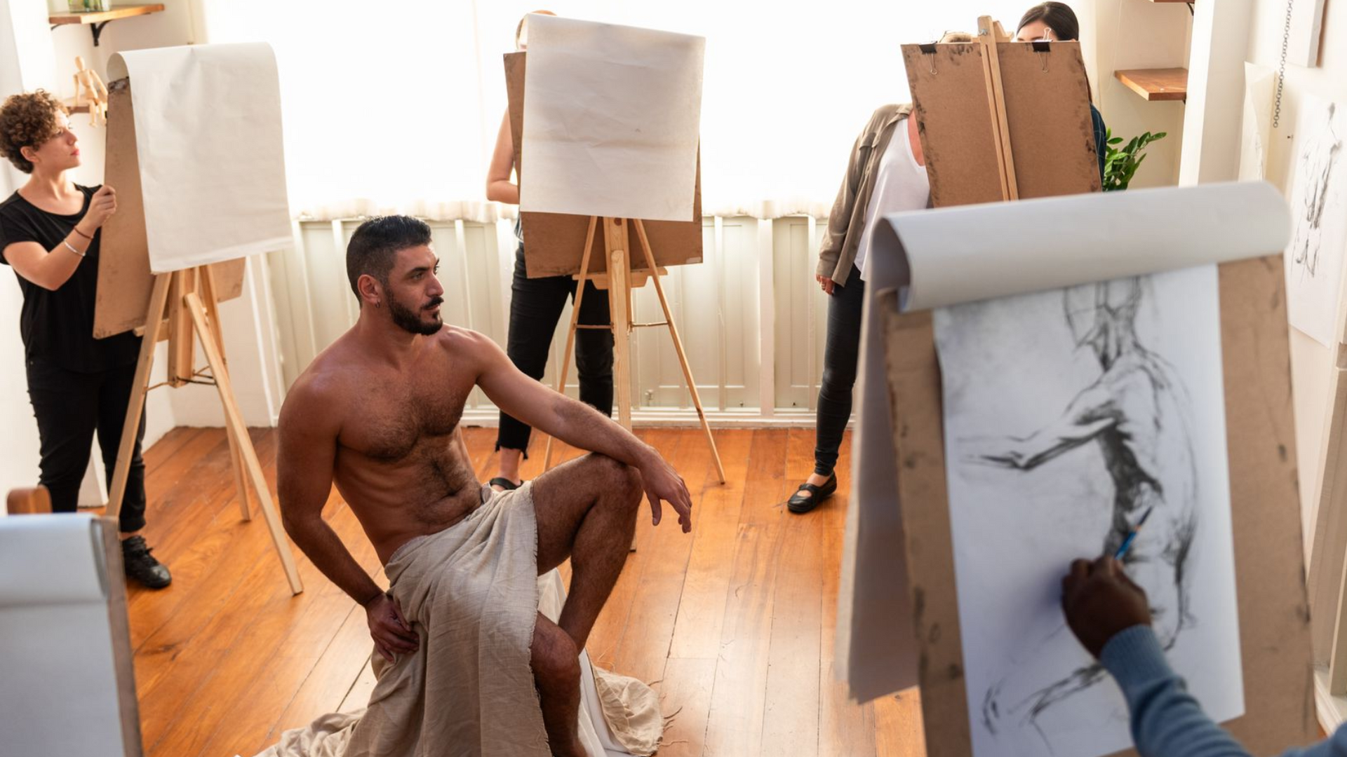 Art class with nude model posing on a stool while artists sketch at easels in a bright studio