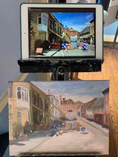 A screen shows a street scene above a painted version of the same view on paper below.