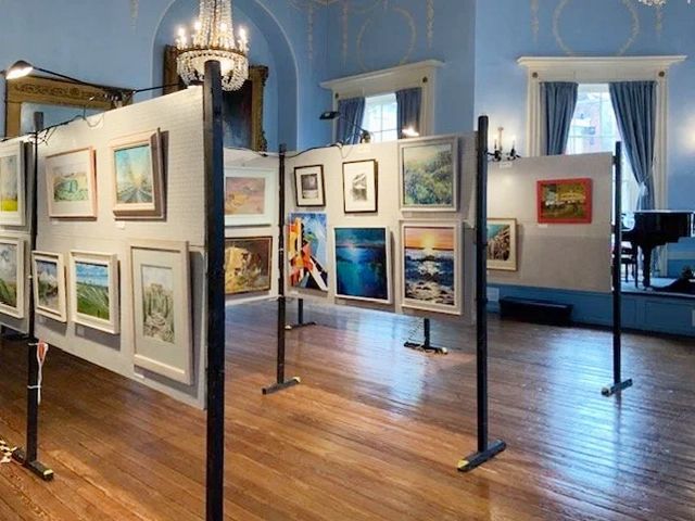 Art gallery interior with framed paintings displayed on freestanding white panels in a blue-walled room.