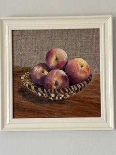 Four apples in a woven basket on a wooden surface, displayed in a framed painting.