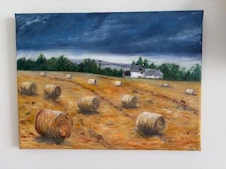 Hay bales in a golden field under a dark cloudy sky, with farm buildings in the distance.