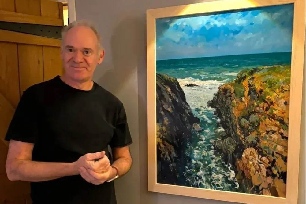 Man standing beside a framed seaside painting with blue sky, ocean, and rocky cliffs
