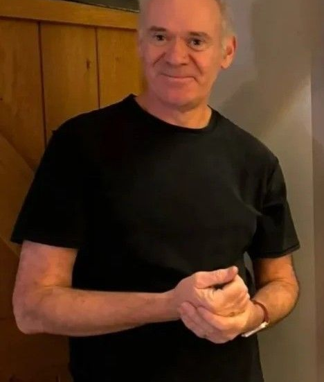 Man in a black T-shirt standing indoors, smiling with hands clasped.