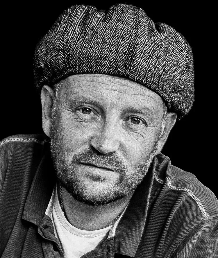 Black-and-white portrait of a man wearing a textured cap and collared shirt, looking at the camera.