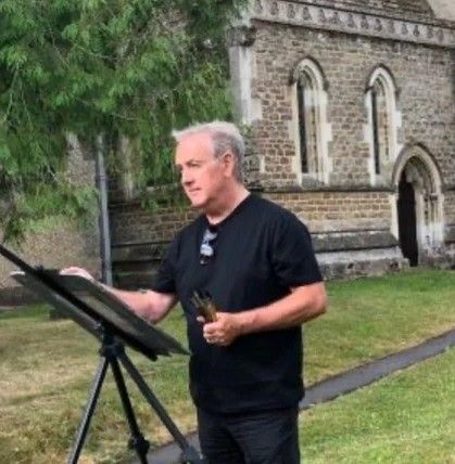 Man standing outdoors beside an easel, holding a brush and painting near a stone building and trees