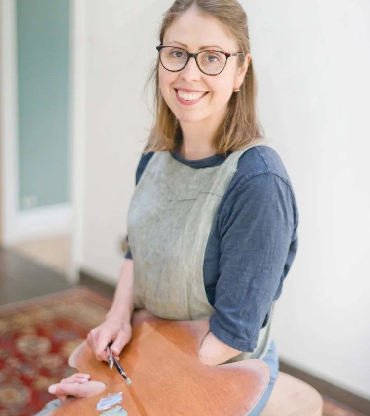 Smiling person in glasses and apron, painting a clay pot at a table indoors