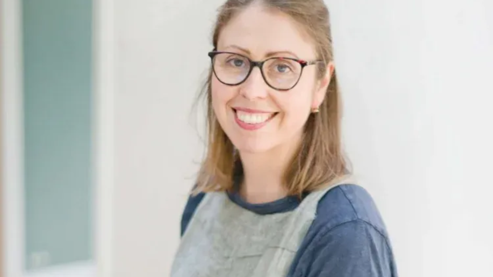 Smiling person with glasses and long hair in a gray top against a light background