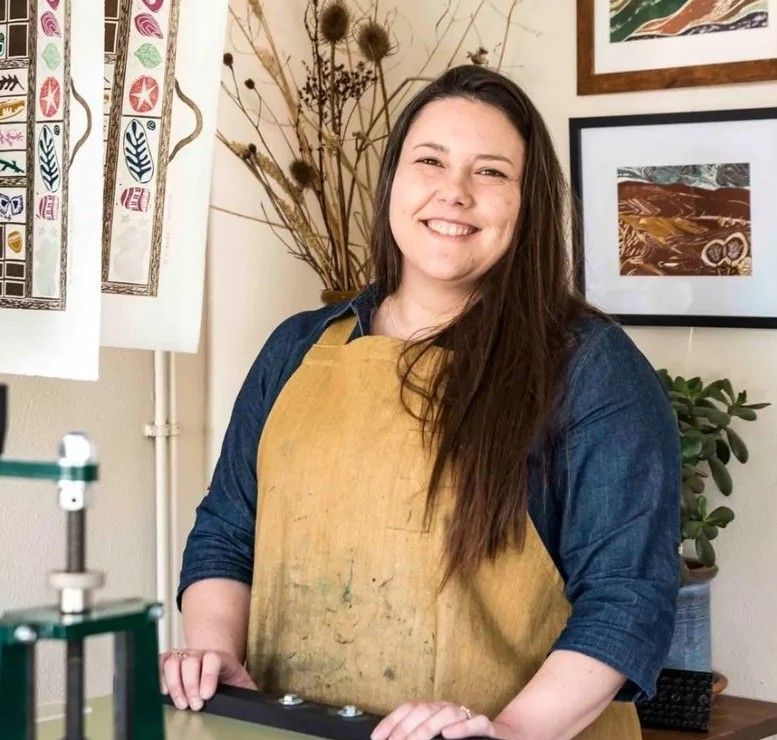 Smiling artist in a blue shirt and yellow apron in a studio with framed artwork and dried flowers