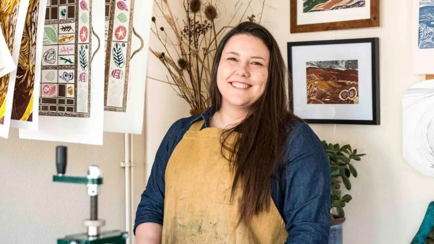 Smiling person in a yellow apron stands in a bright art studio with hanging prints and framed artwork.