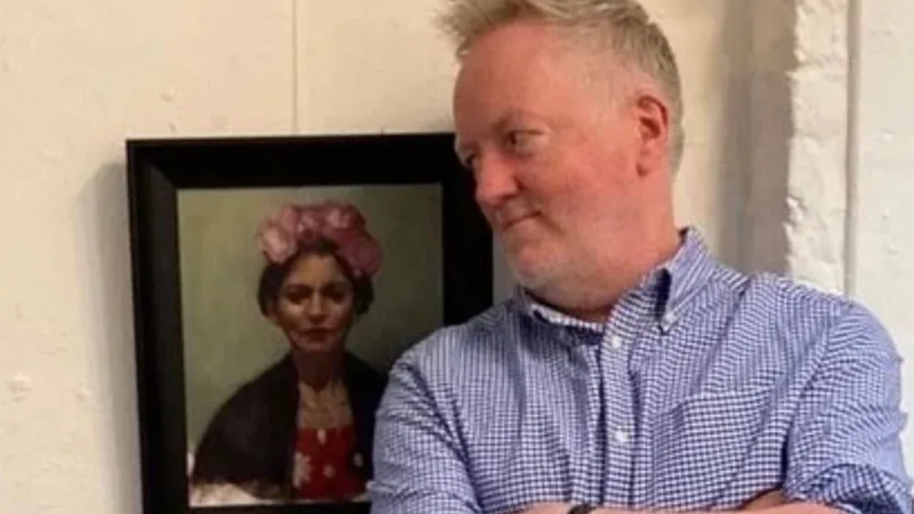 Man standing beside a framed portrait on a wall, wearing a blue checkered shirt and looking to the side