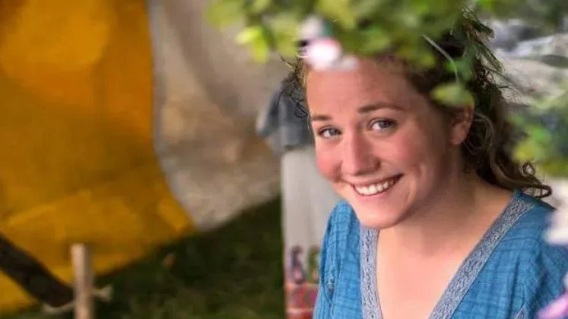 Smiling woman in a blue sweater, partially framed by leaves, outdoors near a yellow tent