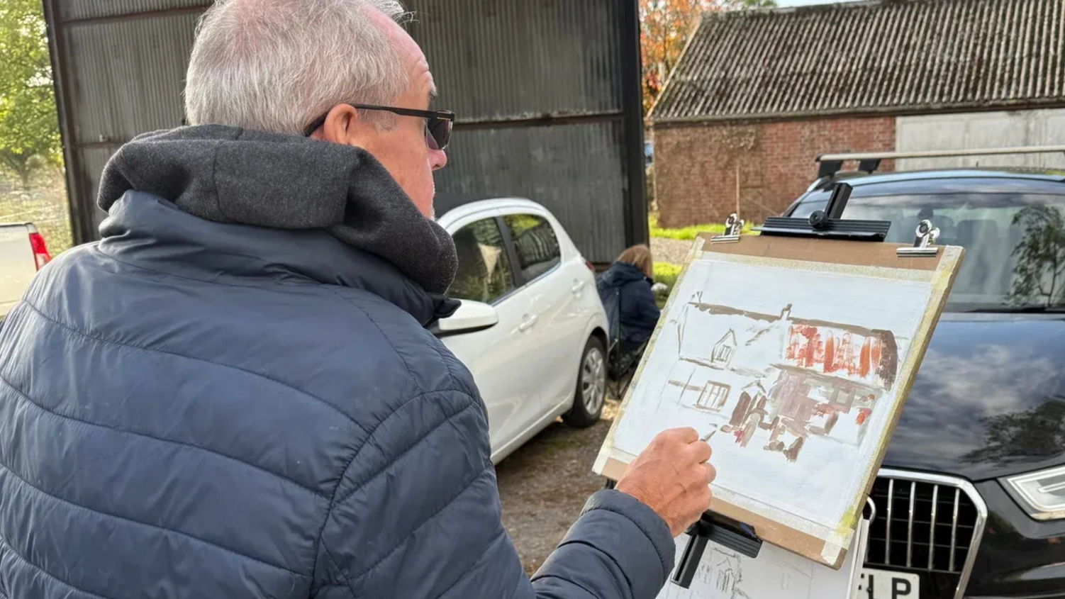 Man in a blue jacket studies a framed print outdoors beside parked cars and a shed.
