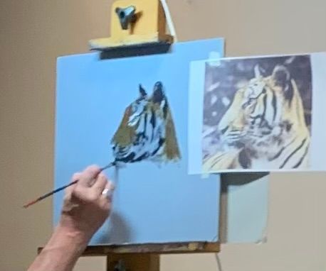 Hand painting a tiger portrait on a blue canvas beside a reference painting