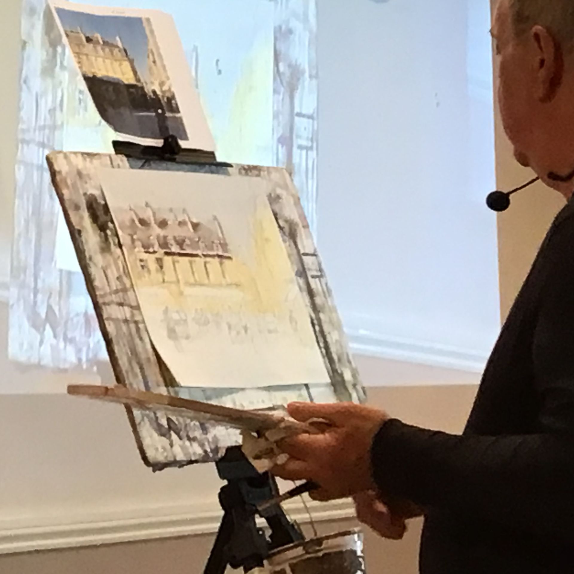Man painting at an easel in a bright studio, working on a sketch with a brush.