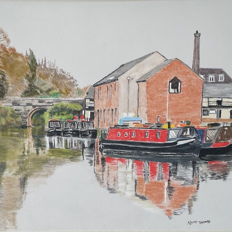 Canal scene with red narrowboats beside brick buildings and a stone bridge, reflected in calm water.