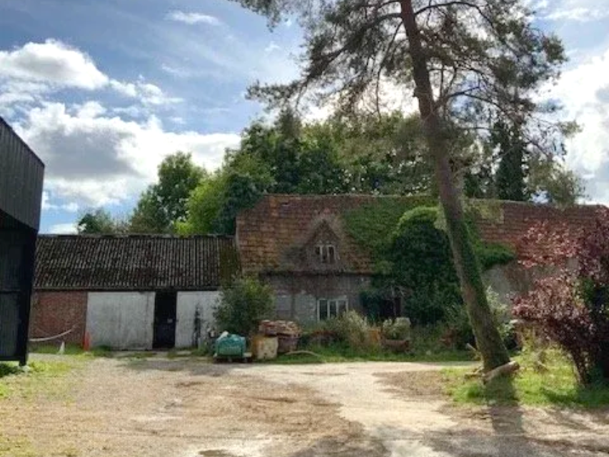 Old rural house with tiled roof, overgrown yard, and a leaning tree under a cloudy sky