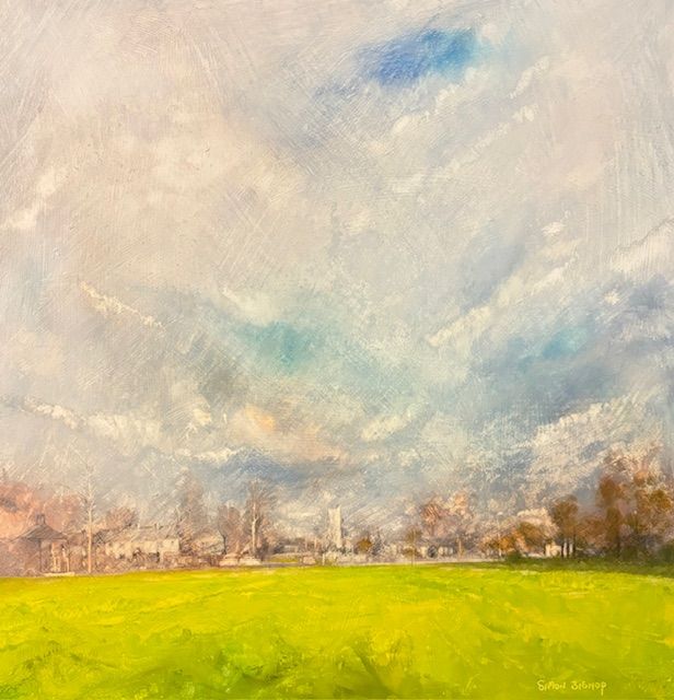 A bright green field beneath a cloudy blue sky with a distant tree-lined town skyline.