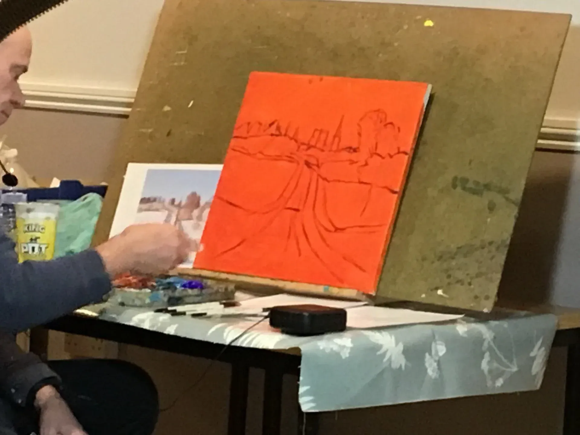 Person painting on an orange canvas at an easel, with a reference photo on the table.