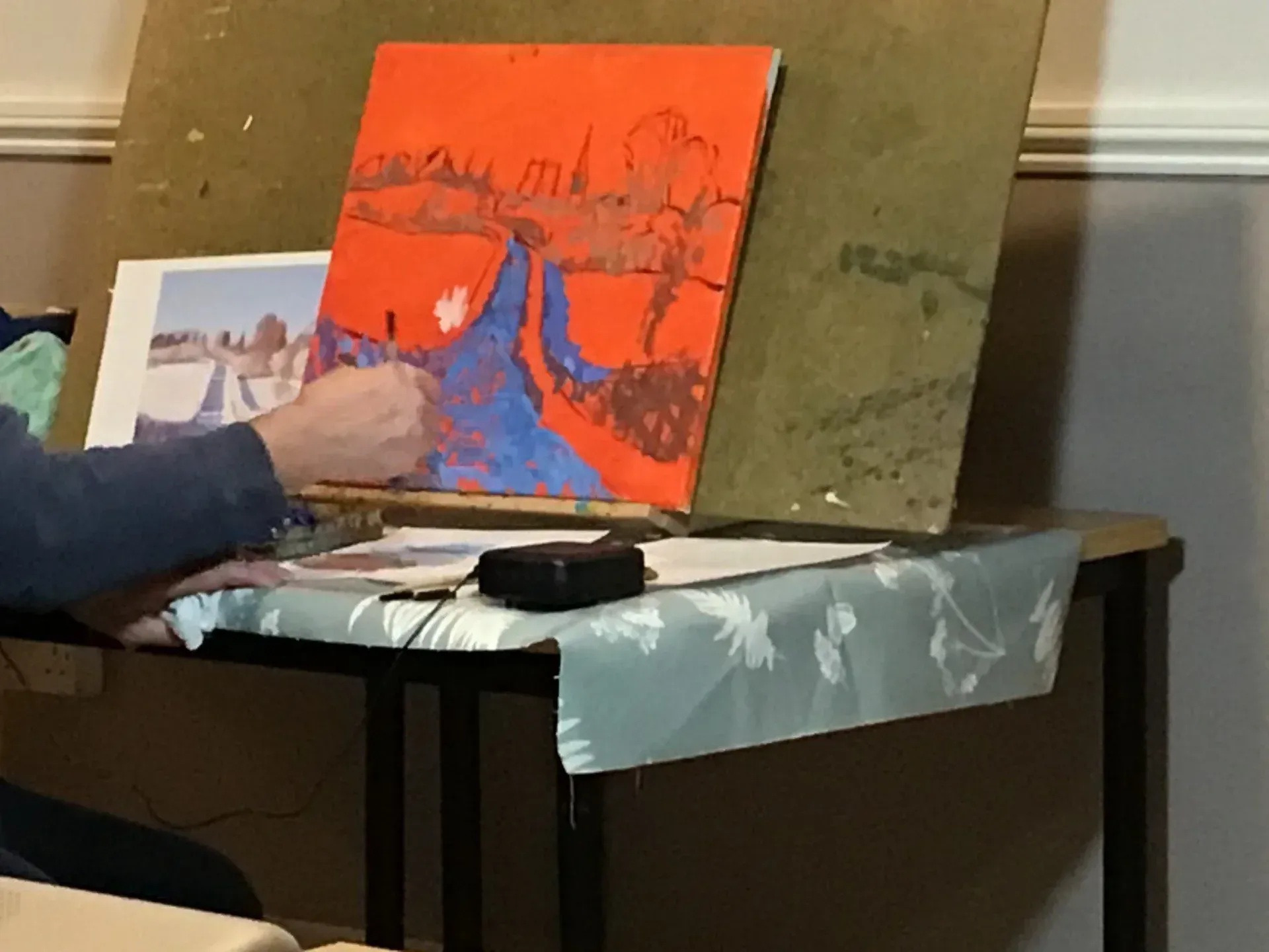 Person painting a bright orange and blue landscape on canvas at an easel in a studio
