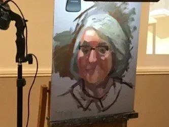 Portrait sketch on an easel, showing a person with gray hair and glasses in a studio setting