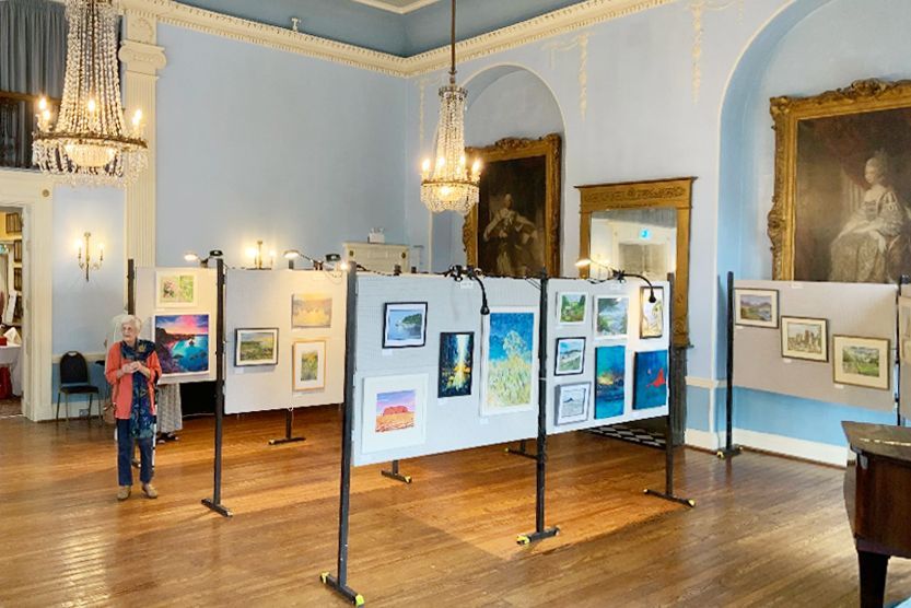 Art gallery with framed paintings on display, chandeliers overhead, and a person viewing the exhibit.