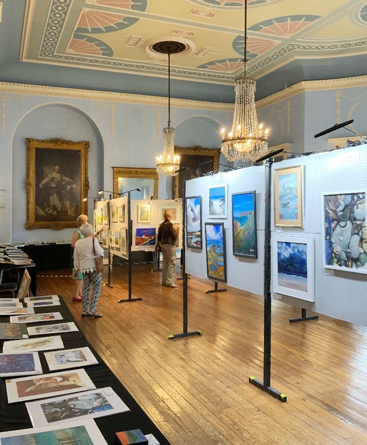 Art gallery exhibition with framed paintings on white walls, chandeliers, and visitors on a polished wooden floor.