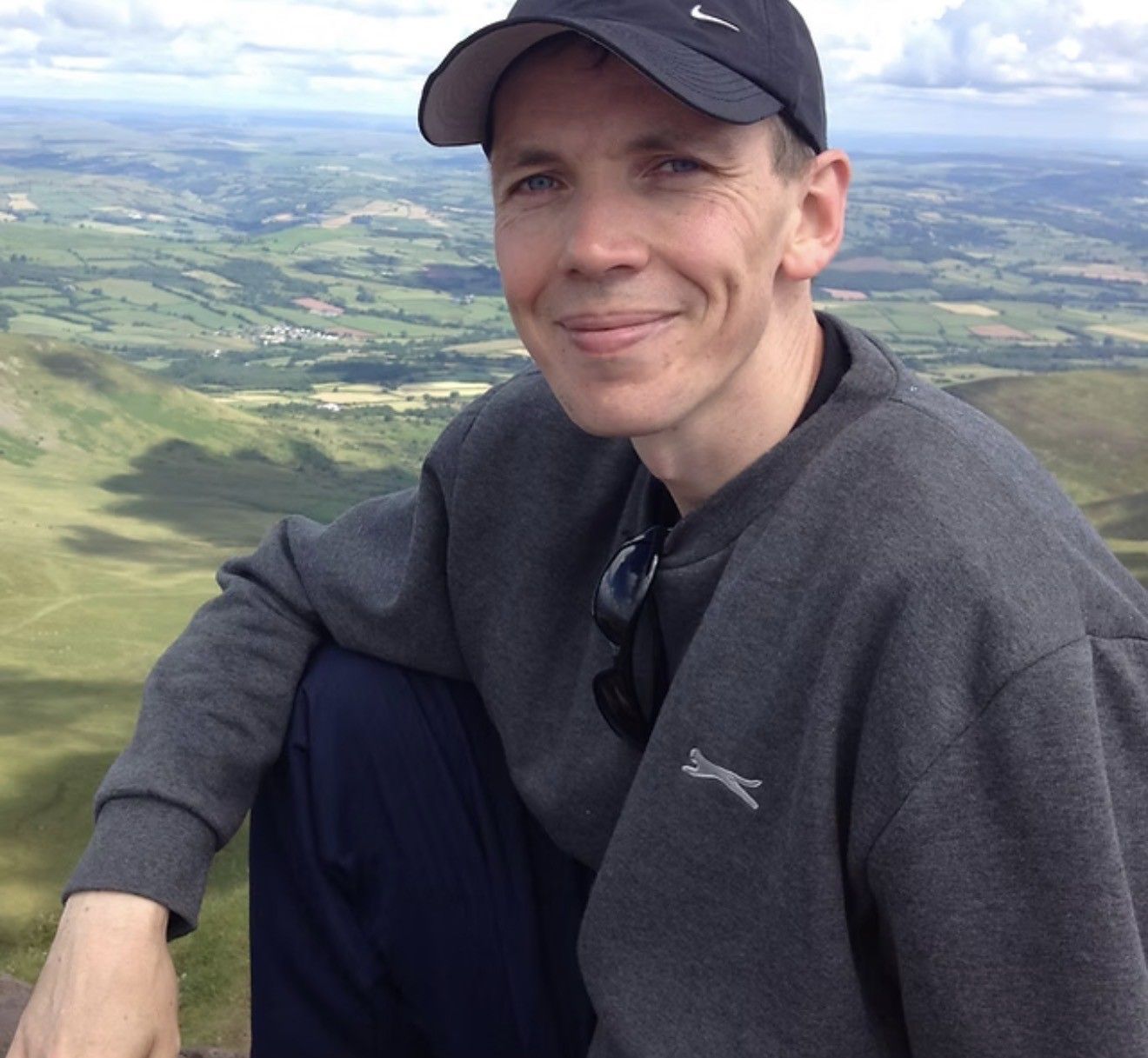 Person in a black cap and gray sweater smiling on a hilltop overlook above a green valley