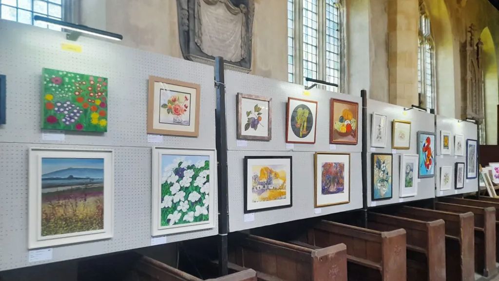 Art exhibition in a long hall, with colorful framed artworks displayed along a wall under large windows.