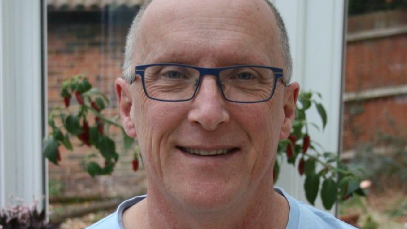 Smiling bald man with glasses in front of leafy plants and a brick wall