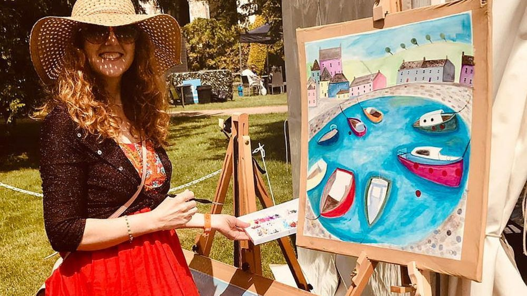 Woman in a hat painting a colorful seaside scene on an easel outdoors.