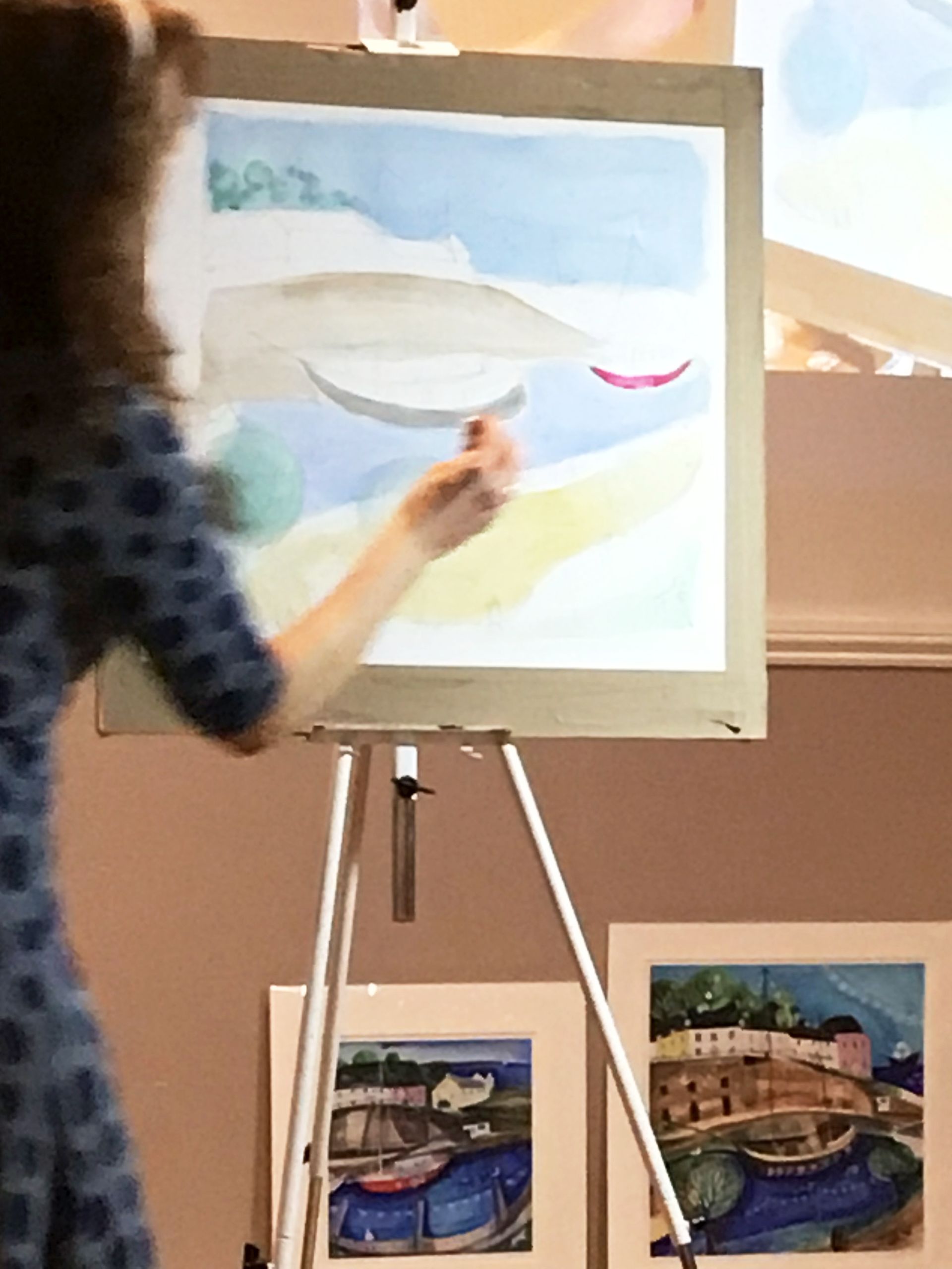 Artist painting a seascape on an easel with two finished landscape paintings nearby
