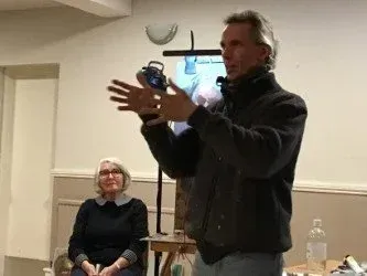 Two people in a room, one speaking and gesturing while standing beside a table and tripod light