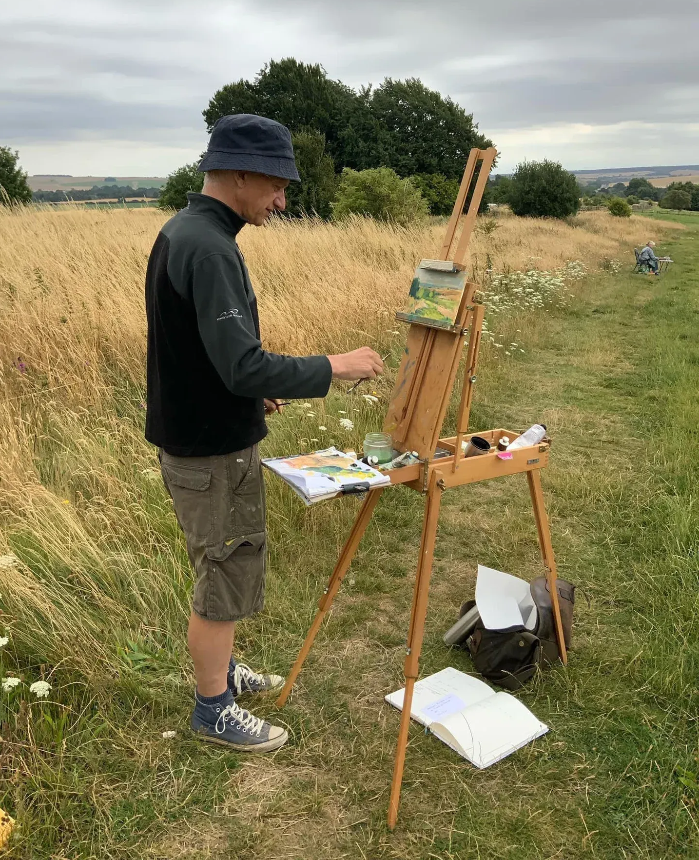 Person painting at an easel in a grassy field beside tall wheat under a cloudy sky