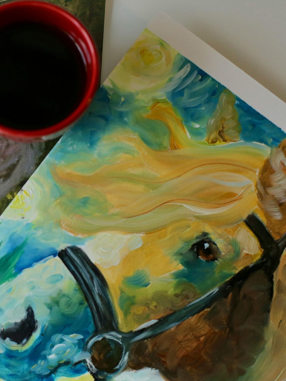 Painted horse face in blue, gold, and brown beside a red cup on a tabletop