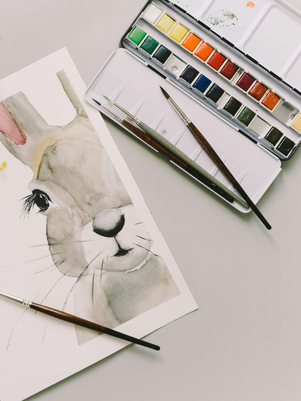 Watercolor painting of a rabbit beside a paint set and brushes on a white table