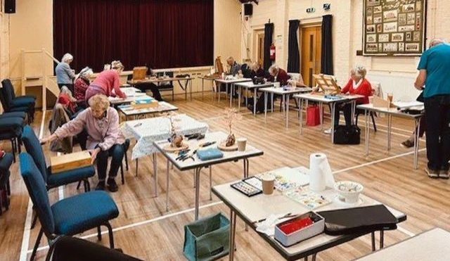 Community room with people seated at tables doing crafts or paperwork, with supplies spread across the floor.