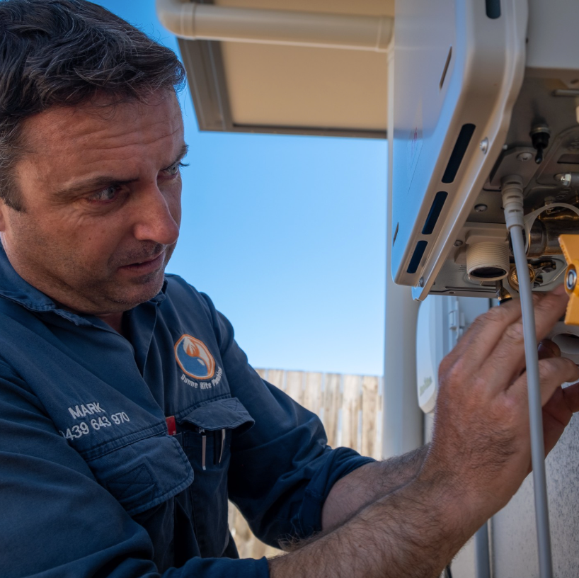 Technician inspecting a hot water system, wearing a blue work shirt outdoors under clear sky — Dunnerite Plumbing in Mount Peter, QLD