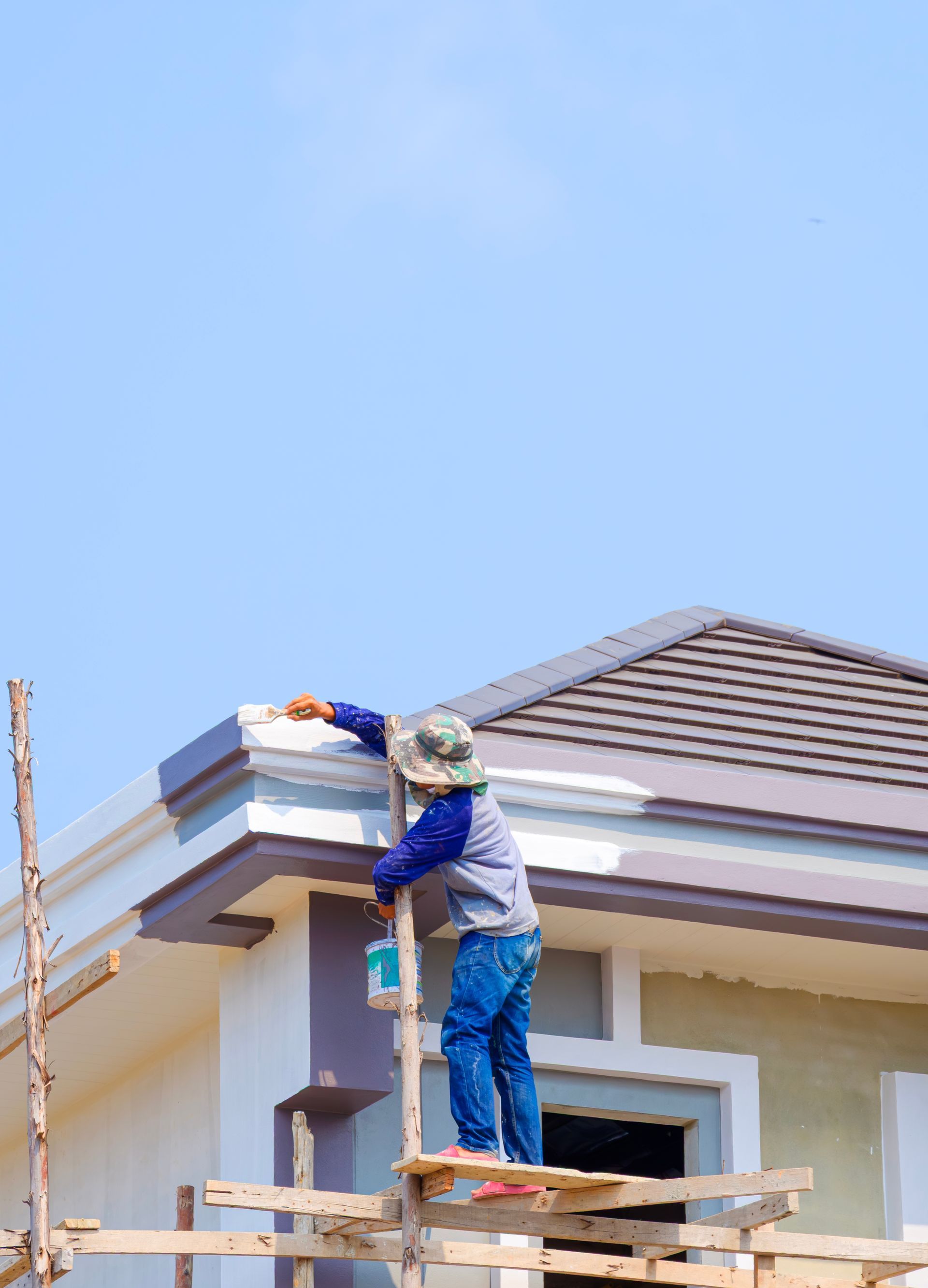Worker painting the exterior trim of a house roof on scaffolding under a clear sky
