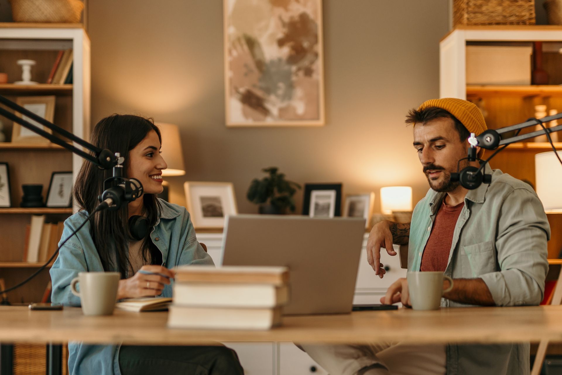 Two people recording a podcast at a table with microphones, laptop, and books in a cozy room