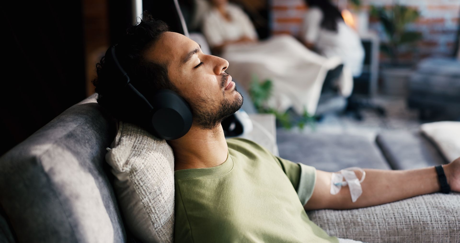 Man reclining on a couch wearing headphones, resting with an IV in his arm.
