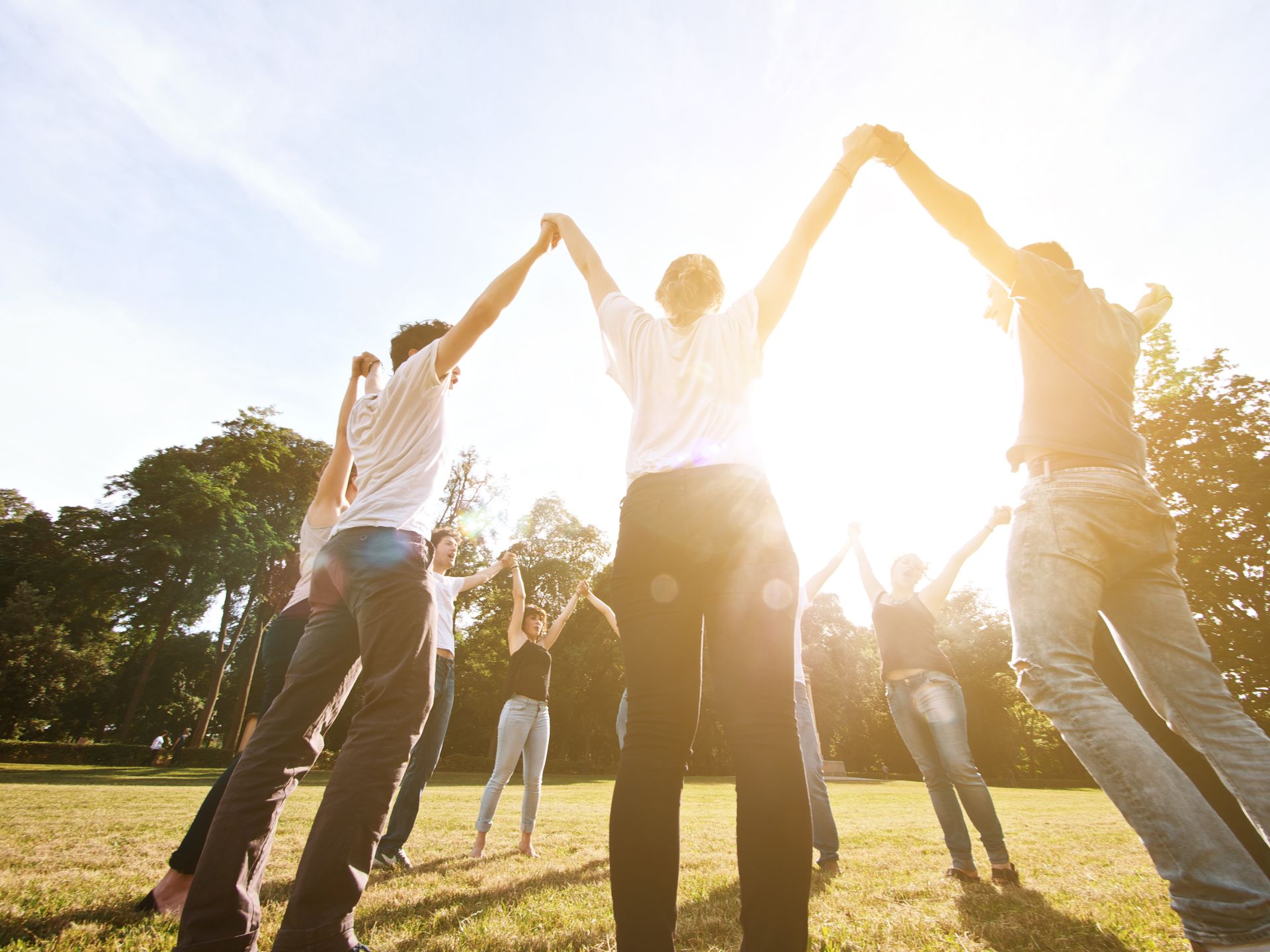 People holding hands in a circle outdoors at sunset, backlit by bright sunlight.