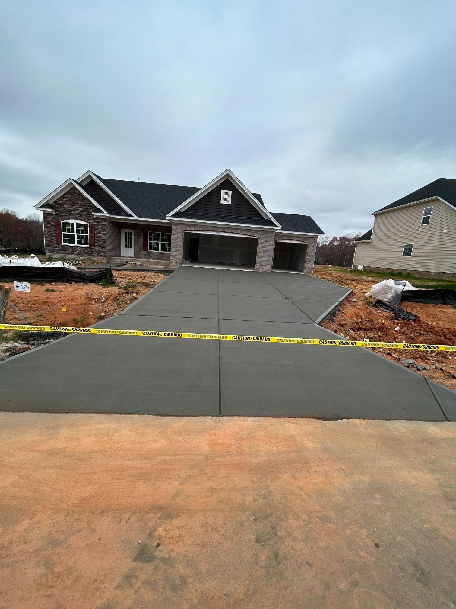 New house with a recently poured concrete driveway, gray on brown earth, under a cloudy sky.