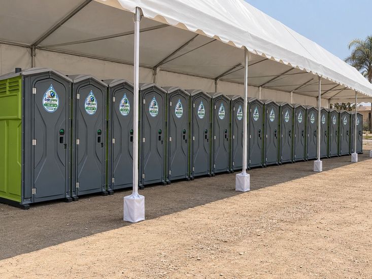 Fila de baños portátiles grises bajo una carpa blanca en un evento al aire libre.