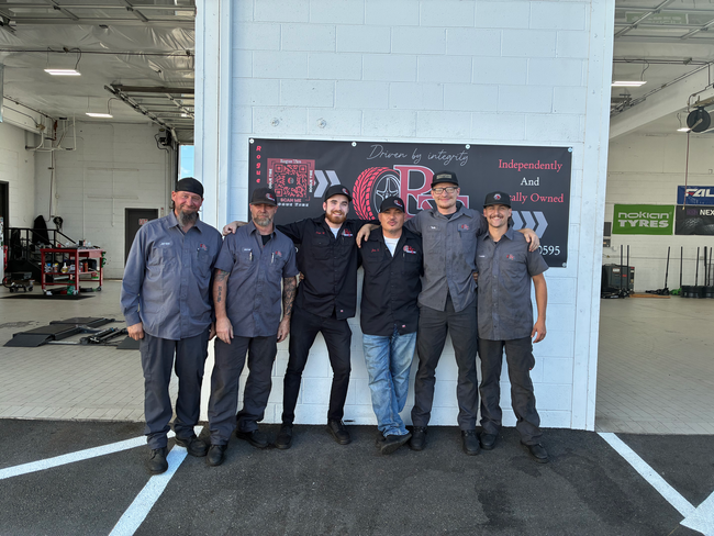 Six people stand together in front of a white brick wall and event banner in a hangar.