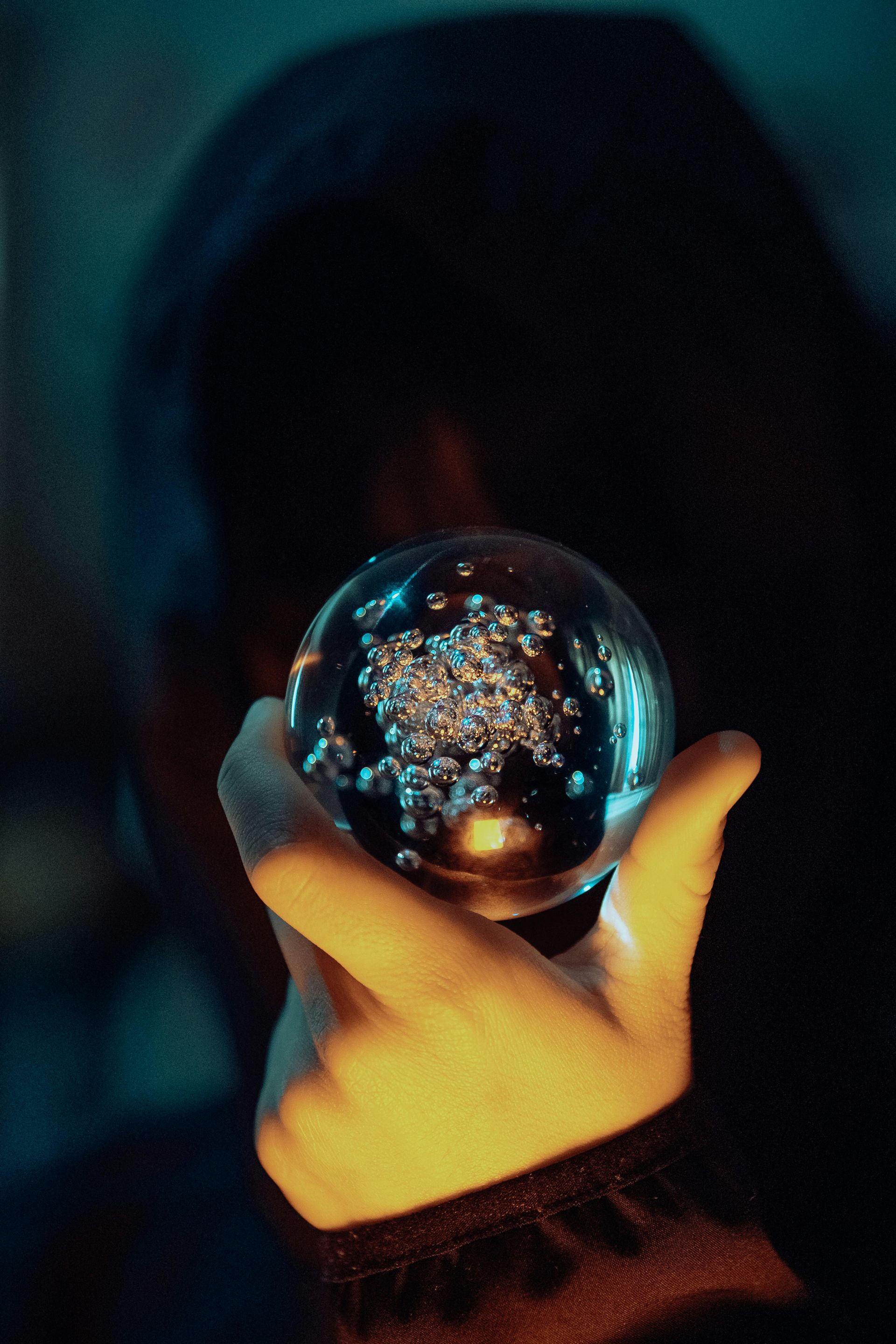 Hand holding a crystal ball filled with sparkling bubbles; person in dark hood obscured in shadows.