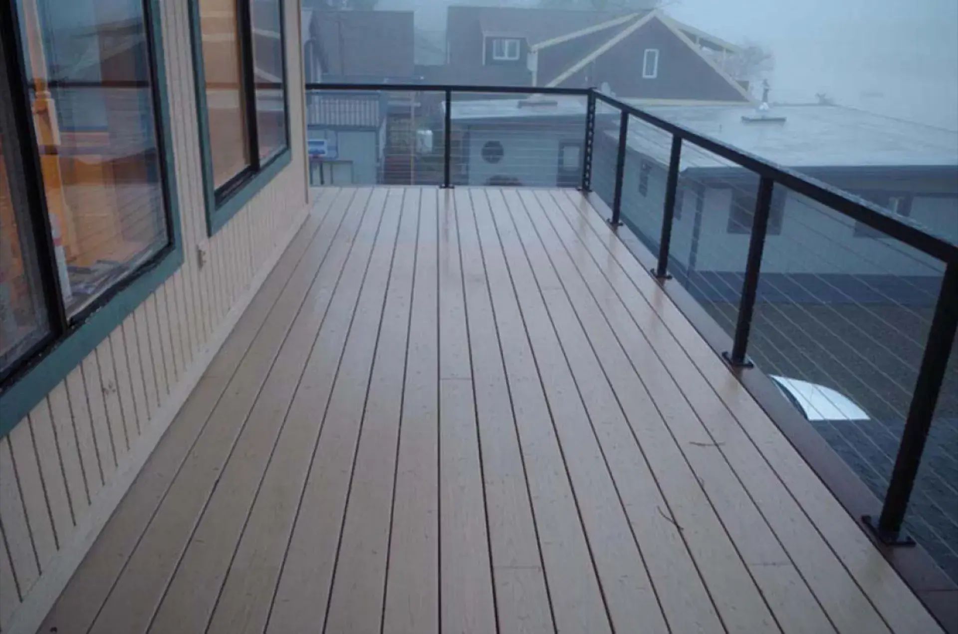 Foggy rooftop balcony with wood decking, glass railings, and a house in the distance