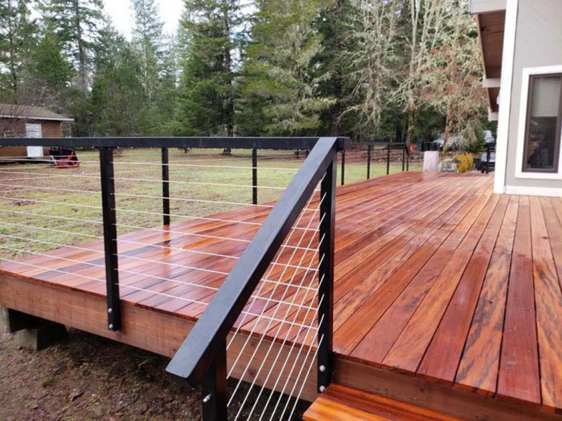 Wooden deck with black cable railing and wet reddish boards overlooking a yard and trees