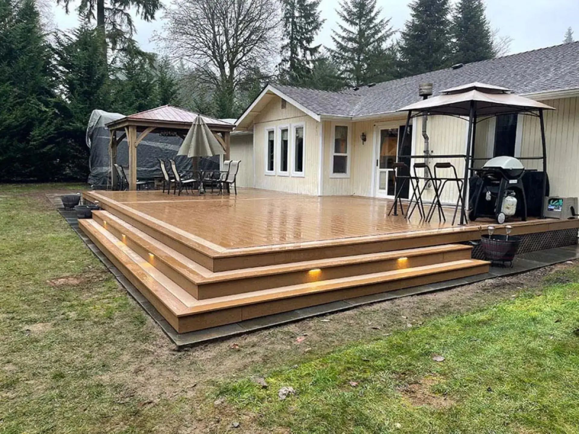Freshly built wooden deck beside a beige house, with outdoor furniture and a grill in the yard